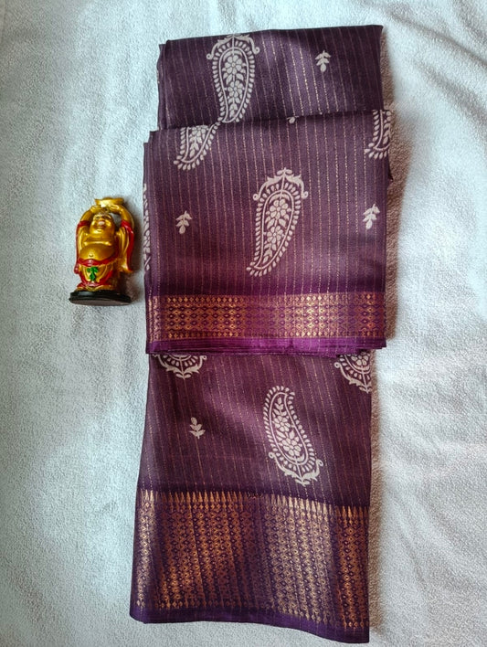 A folded purple silk saree with white paisley motifs and a metallic gold border, displayed on a white textured fabric background next to a small gold figurine.